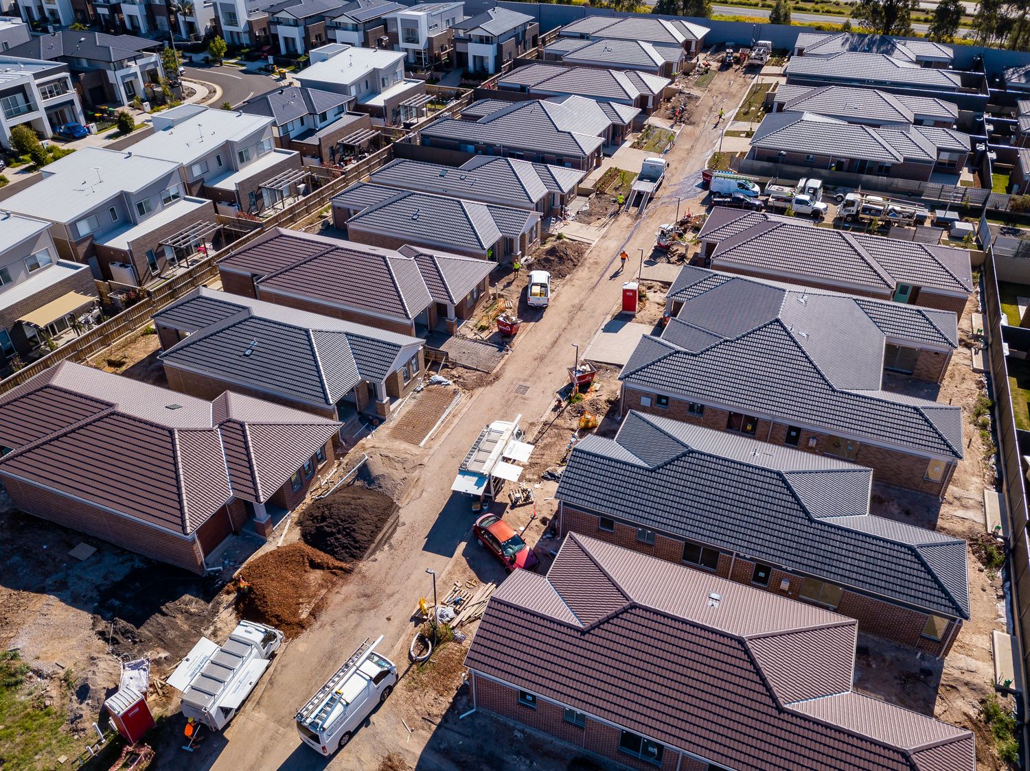 Drone photo of a housing estate under construction showing brick houses, tiled roofs, construction vehicles, and unpaved roads.