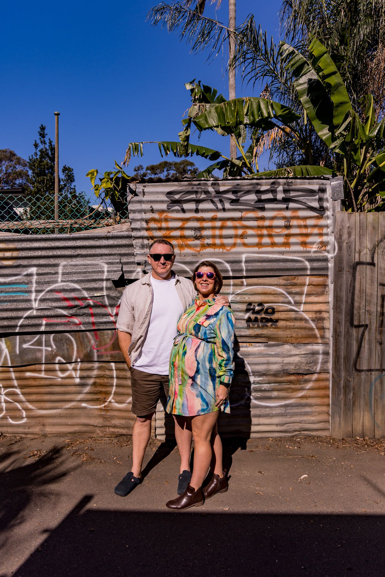 Smiling couple in sunglasses embrace in front of a graffiti-covered corrugated metal fence with tropical plants behind them.