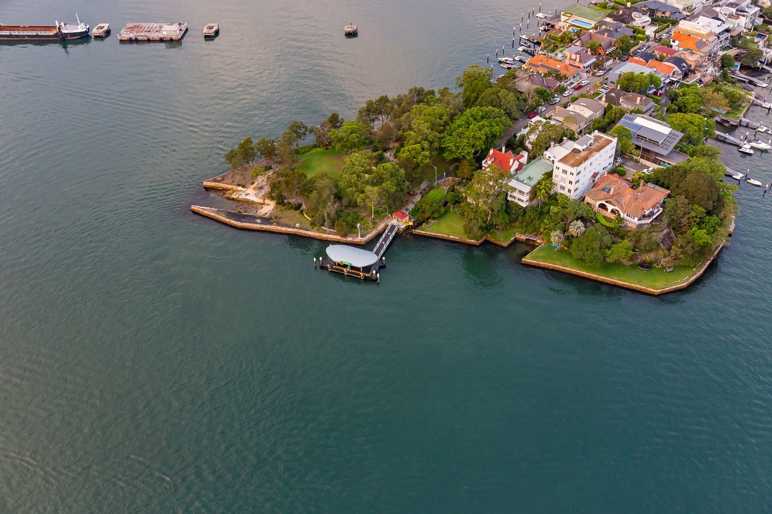 Aerial view of a ferry wharf on a leafy harbour peninsula with red-roofed houses, green parkland and deep teal water.