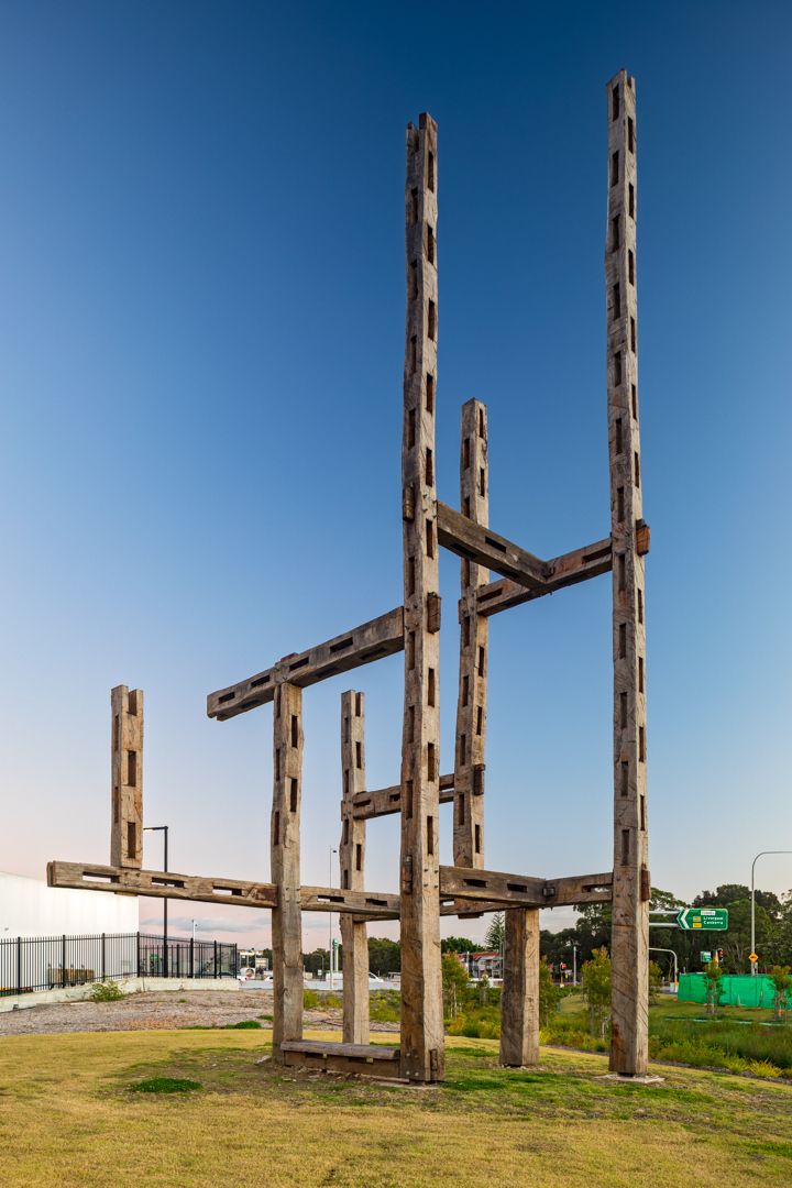 Tall weathered timber sculpture with crossbeams rising from a grassy lawn against a clear blue dusk sky.