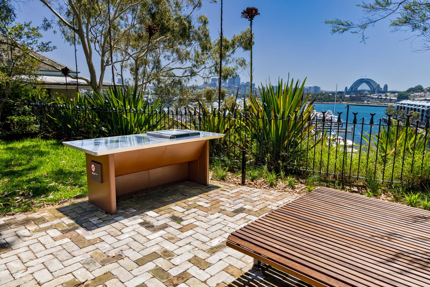 Paved garden terrace with a modern outdoor barbecue bench, native plants and a black fence overlooking a harbour and distant bridge.