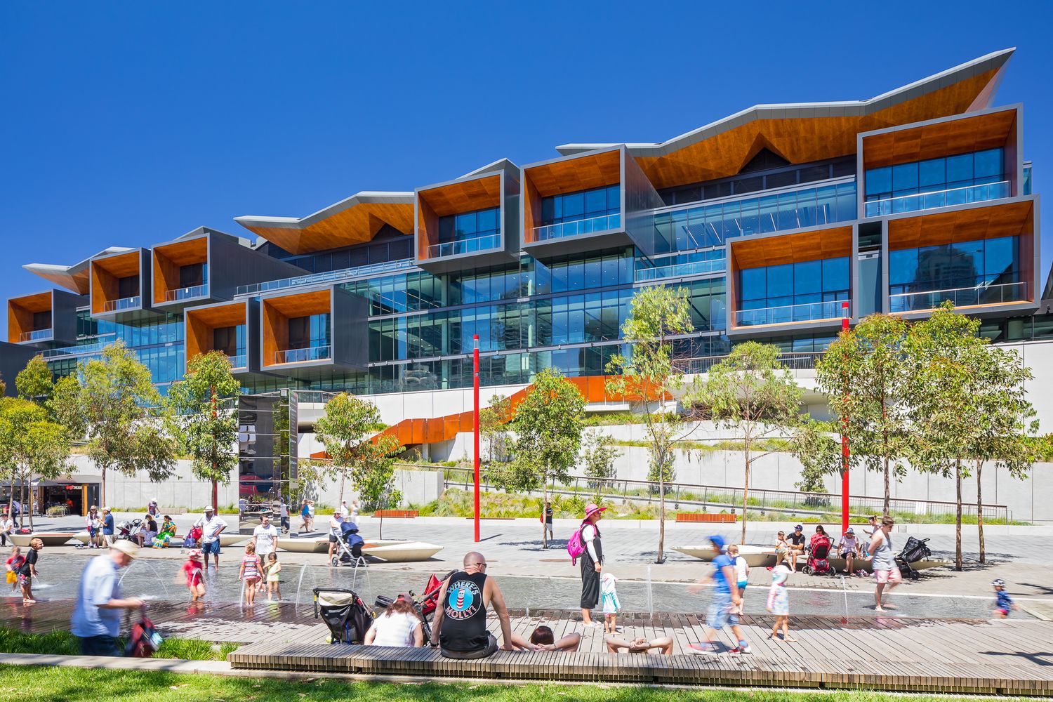 Families relax on a sunny waterfront promenade in front of a modern glass convention centre with orange timber accents.