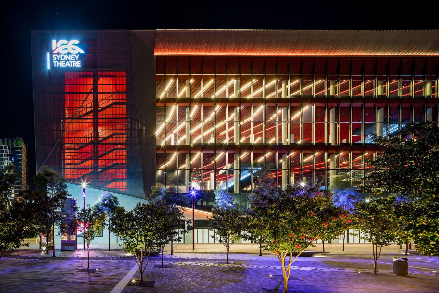 Sydney Theatre building lit up at night with glowing orange and white lighting behind a glass facade and landscaped trees.