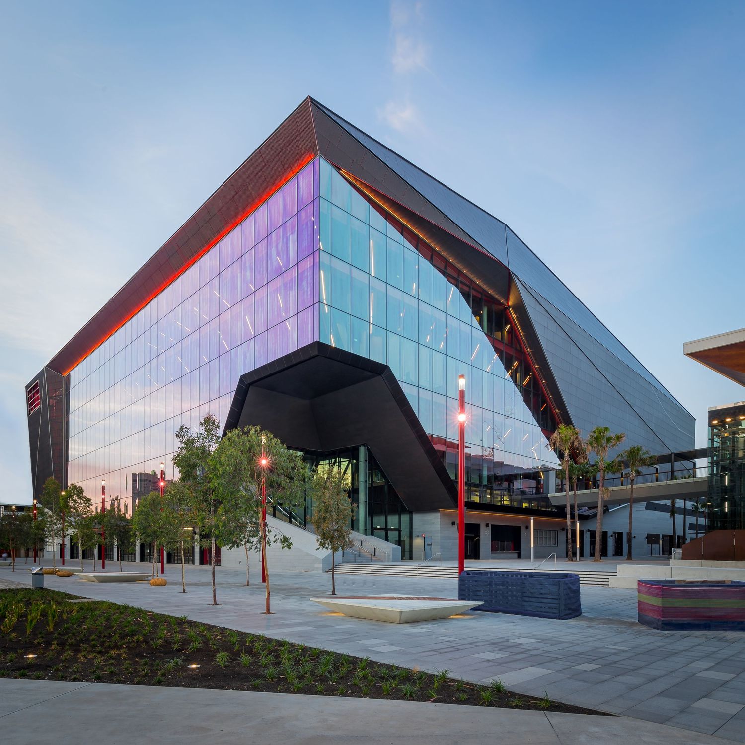 Modern angular glass convention center at dusk with a dark geometric canopy, red accents and trees lining a paved public plaza.