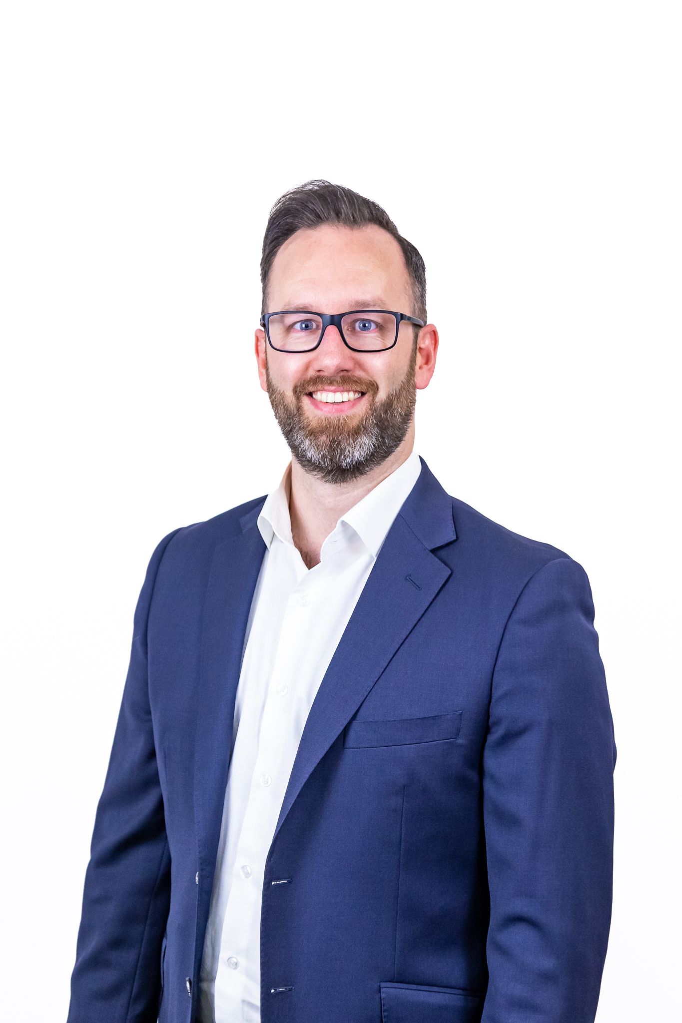 Smiling bearded man in glasses, a navy blazer and white open-collar shirt poses for a professional portrait on a white background.