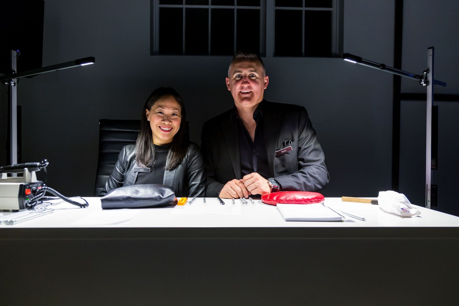 Smiling woman and man in dark clothing sit at a white desk with a lamp, tools and a red dish in a dimly lit studio.