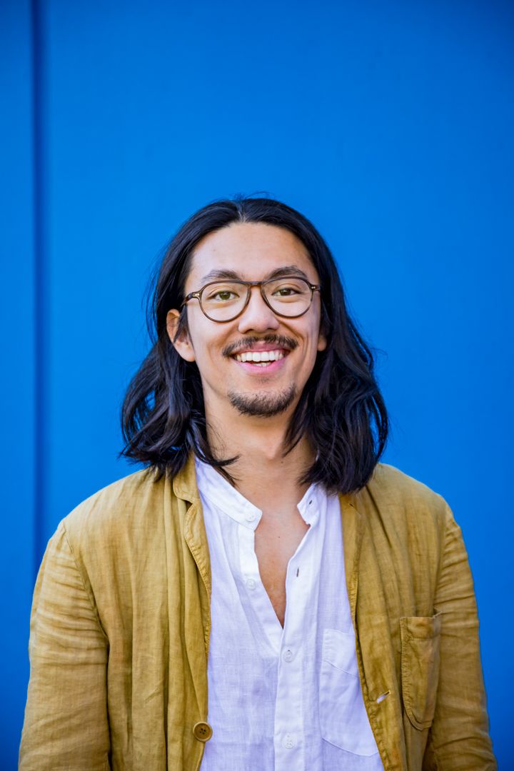 Smiling man with shoulder-length dark hair, round glasses and a goatee wears a mustard linen jacket against a bright blue wall.