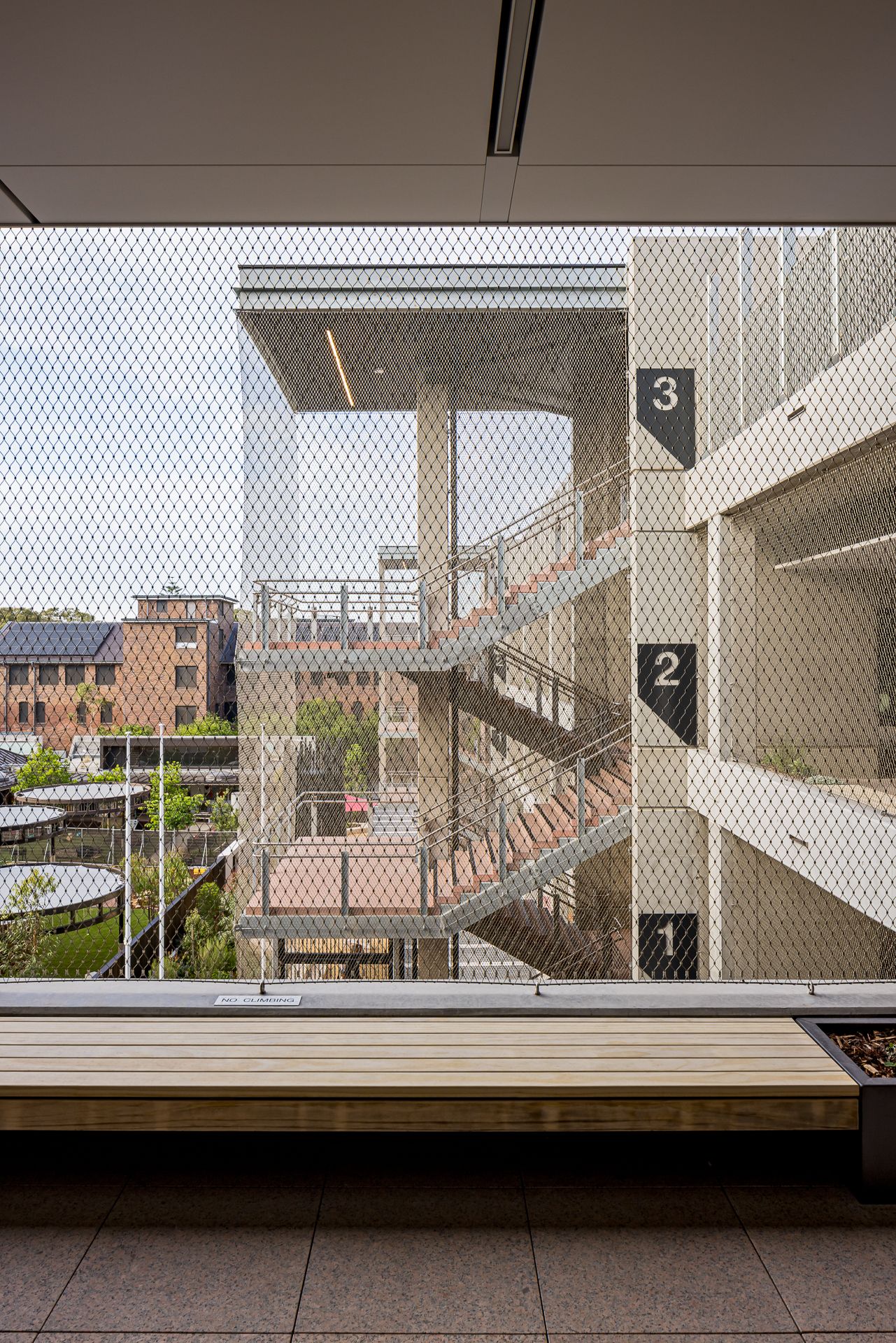 View through a wire mesh screen of an exterior staircase and numbered building levels, with brick buildings and greenery in the background.