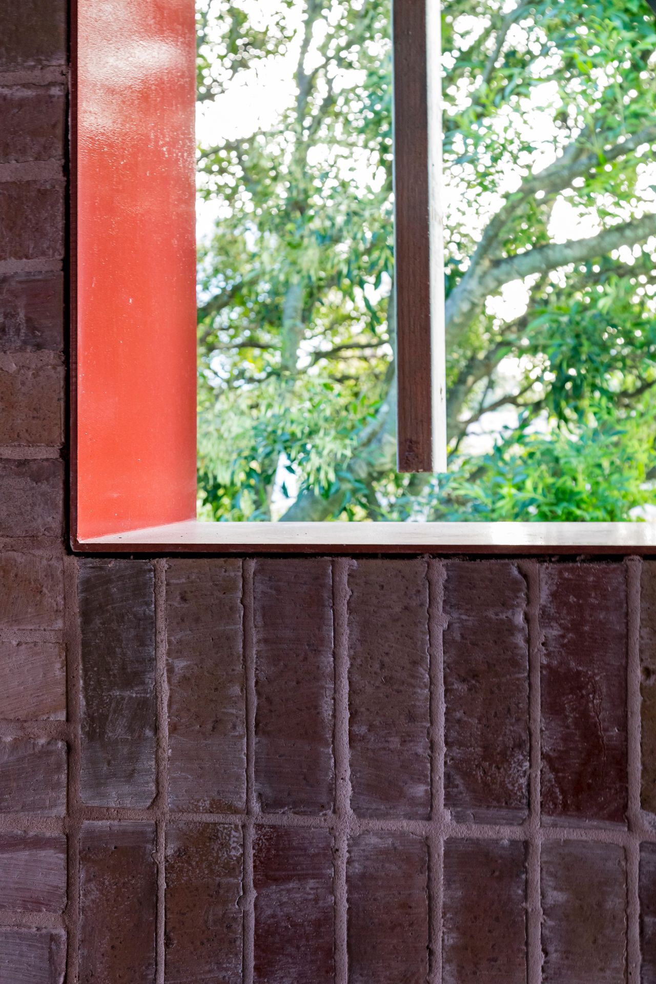 Red-framed window opening in a rustic brick wall looking out onto green leafy tree branches.