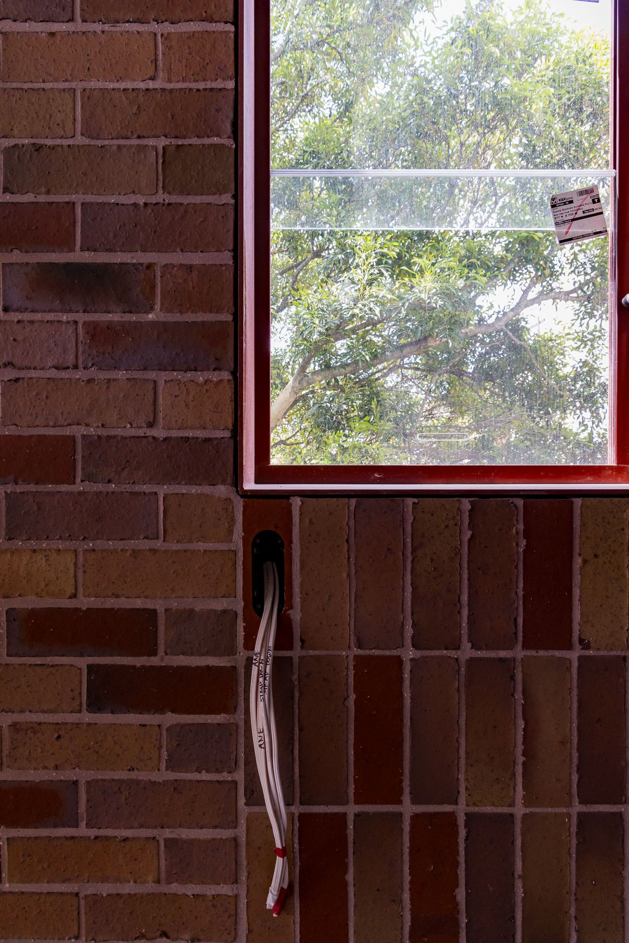Red-framed window showing green tree foliage beside a mix of brown and red brick walls, with loose electrical cable hanging below.
