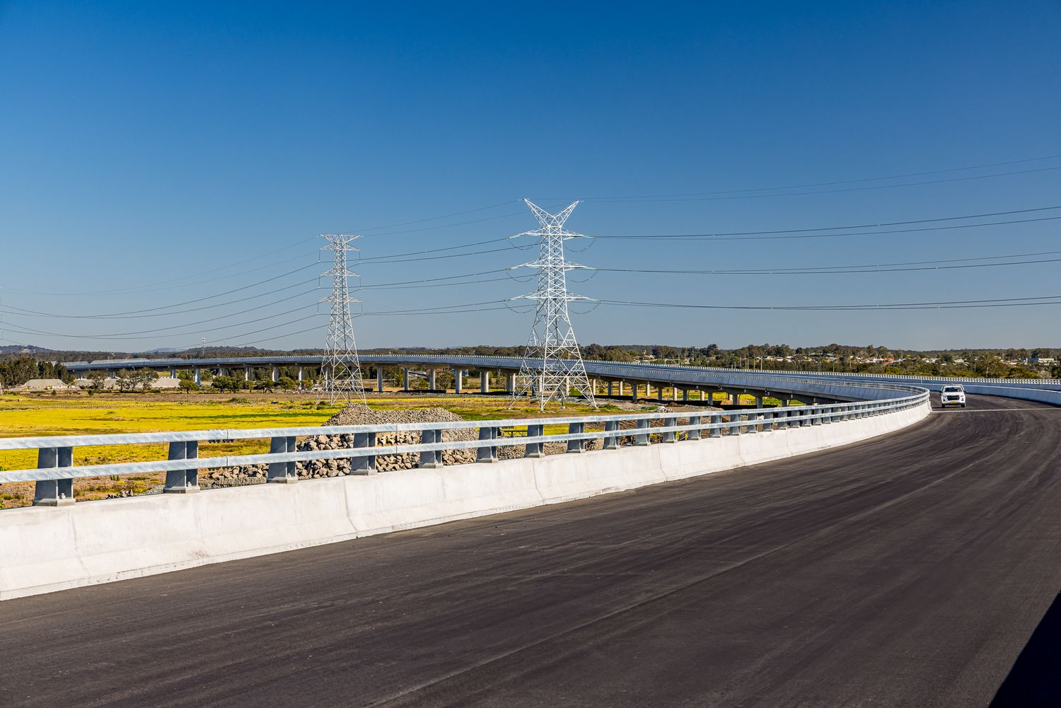 Freshly paved elevated motorway with concrete barriers and tall electricity pylons crossing yellow wetland fields under blue sky.