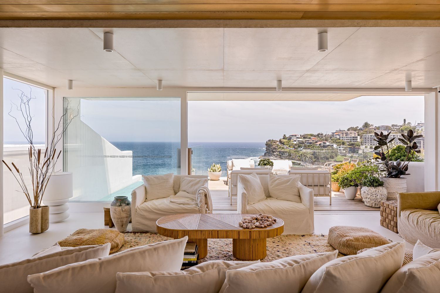 Sunlit living room with white sofas and timber coffee table opening to a terrace with panoramic ocean and headland views.