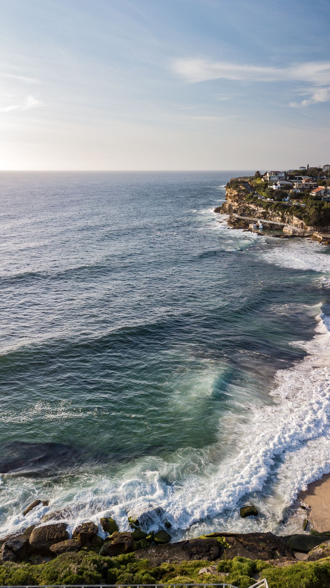 Waves crash against rocky shores along a scenic coastline with clifftop homes in soft evening light.