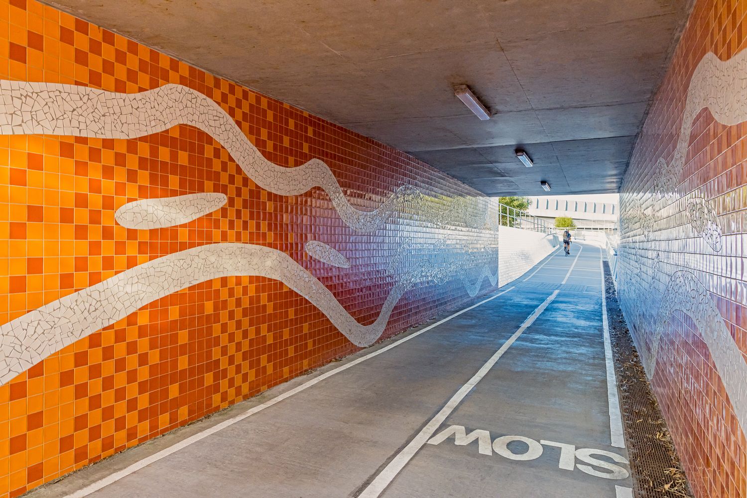 Bright orange tiled underpass with white wave patterns and a bike lane marked slow leading to daylight.