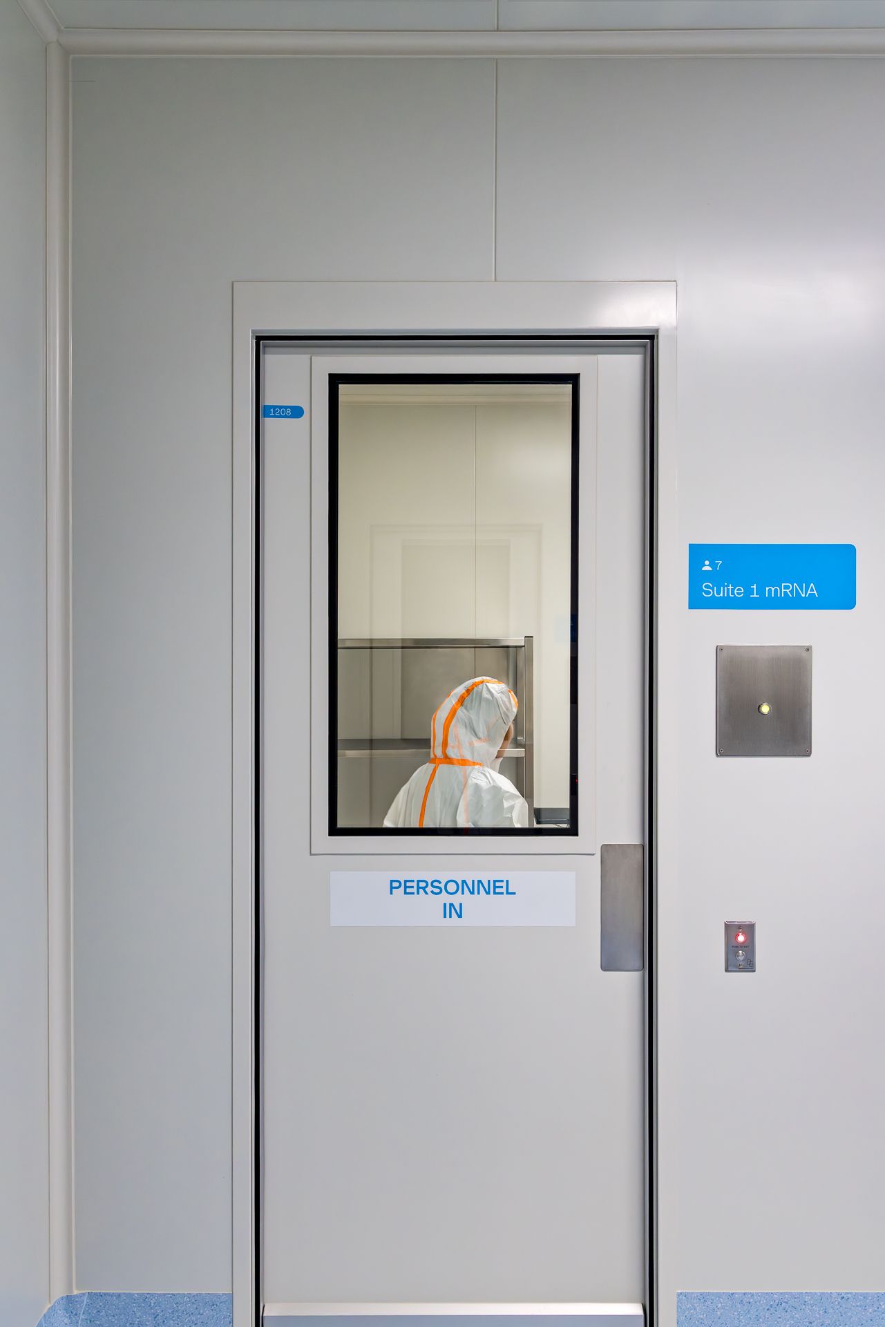 Cleanroom personnel entry door labeled Personnel In for Suite 1 mRNA, with a worker in protective coverall visible through the window.