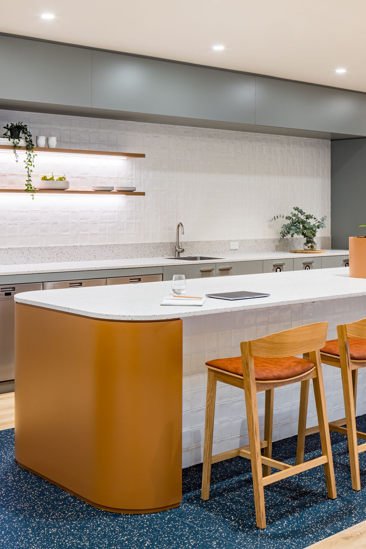 Contemporary office kitchen featuring a curved orange island, white terrazzo benchtop, timber barstools and open shelving with plants.