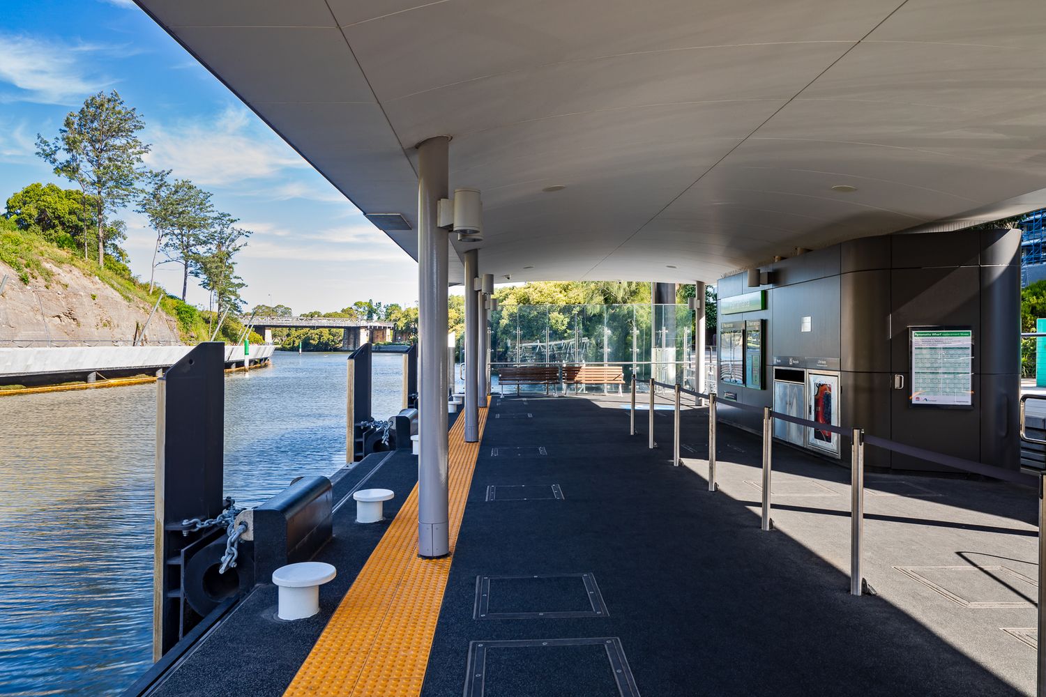 Covered ferry wharf platform with a dark floor, yellow tactile edge and steel railings beside the river and a tree-lined bank.