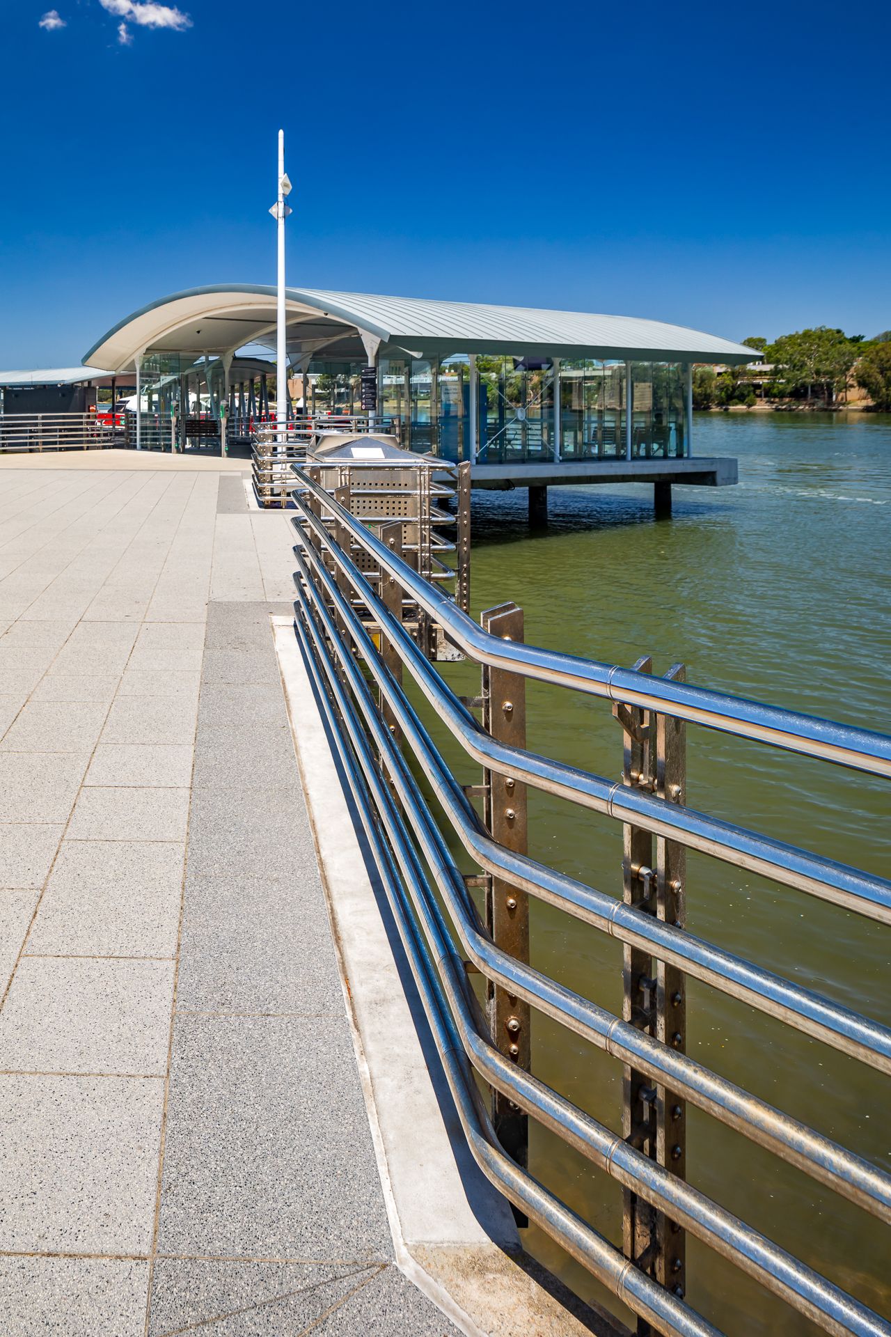 Stainless steel railings and granite paving lead to a covered ferry wharf extending over the water on a bright sunny day.
