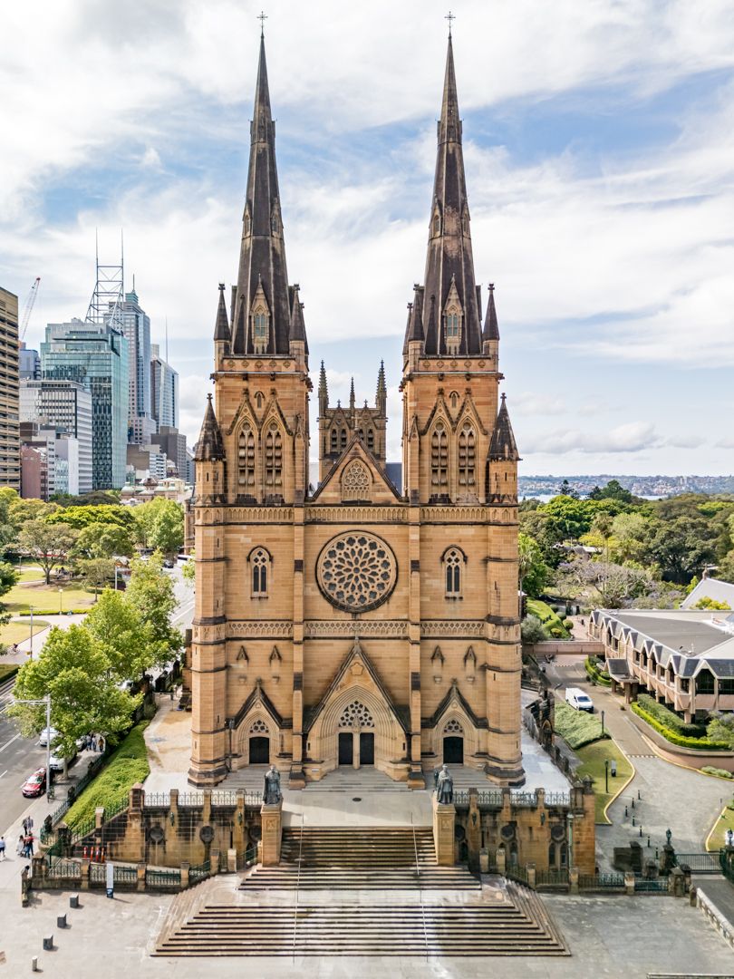 Sandstone Gothic Revival cathedral with twin spires and a rose window, set beside city skyscrapers under a partly cloudy sky.