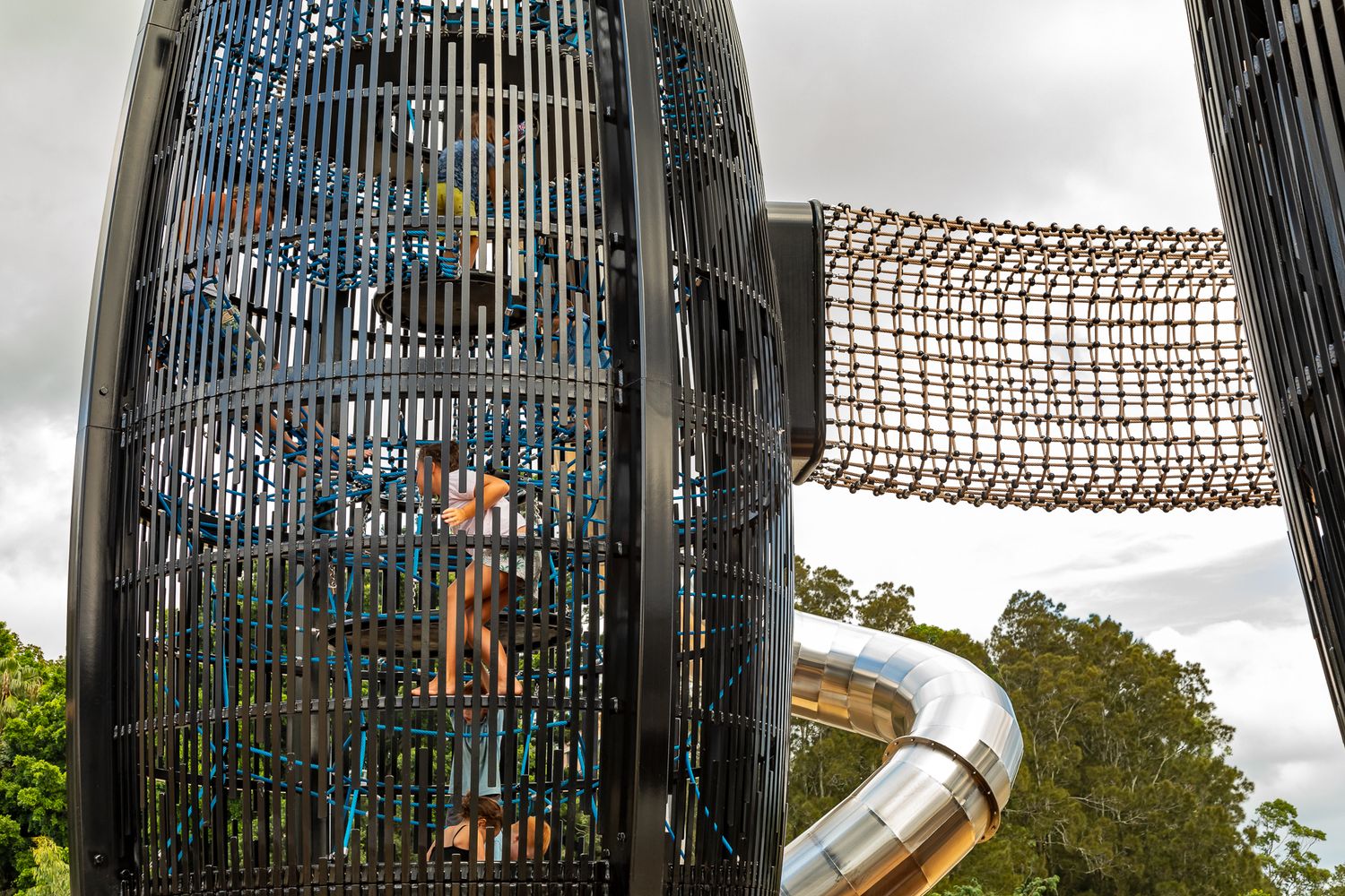 Children climb inside a steel mesh play tower connected by a rope net bridge, with a silver slide curving below.