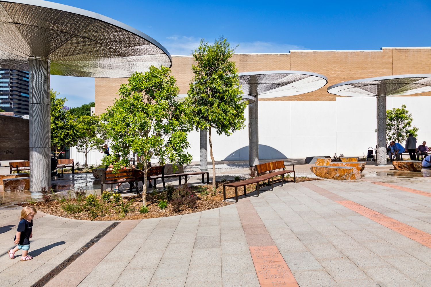 A child walks across patterned paving in a public plaza with young trees, timber benches and canopy shade structures against a blue sky.