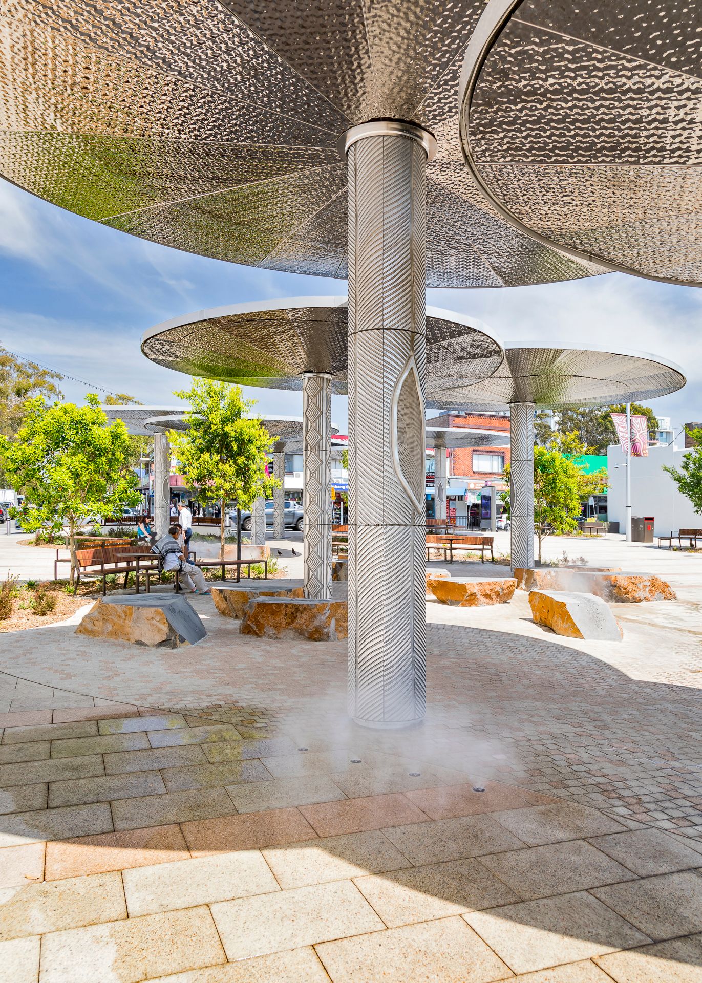 Mist rises from a paved plaza with perforated metal canopy shades, patterned columns, boulder seating and benches.