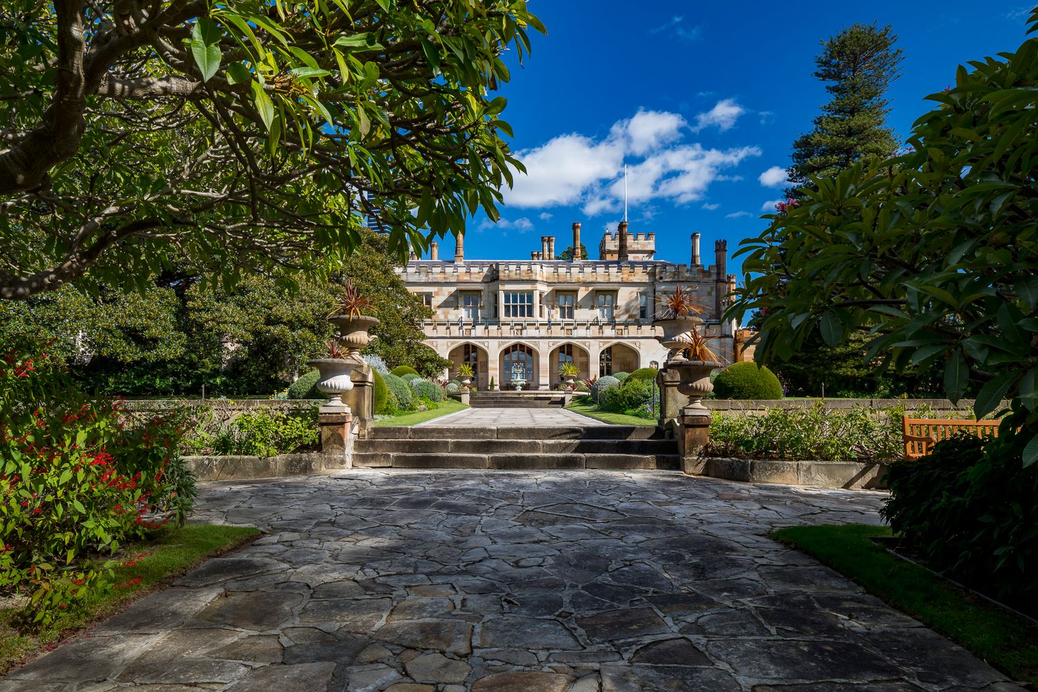 Stone path leads past garden beds and an entrance staircase to a grand Gothic Revival sandstone mansion framed by tall trees.