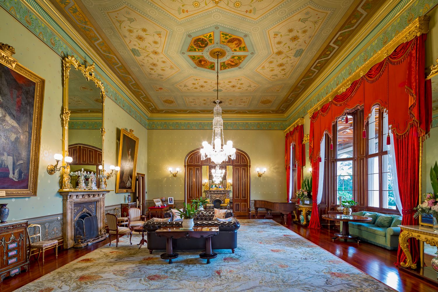Grand historic drawing room with decorative painted ceiling, crystal chandelier, red drapes, fireplace and patterned carpet.