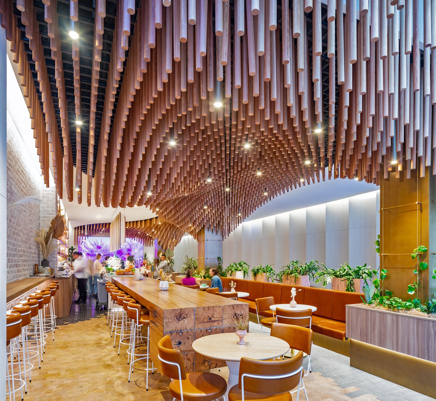 Warm timber ceiling sculpture swoops over a modern cafe with bar stools, round tables, orange chairs and purple flowers.