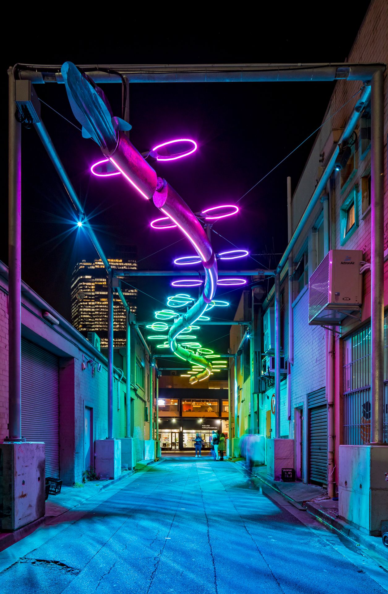 Narrow city laneway at night with a spiral light sculpture overhead glowing in pink, purple and green neon, and a lone passerby.