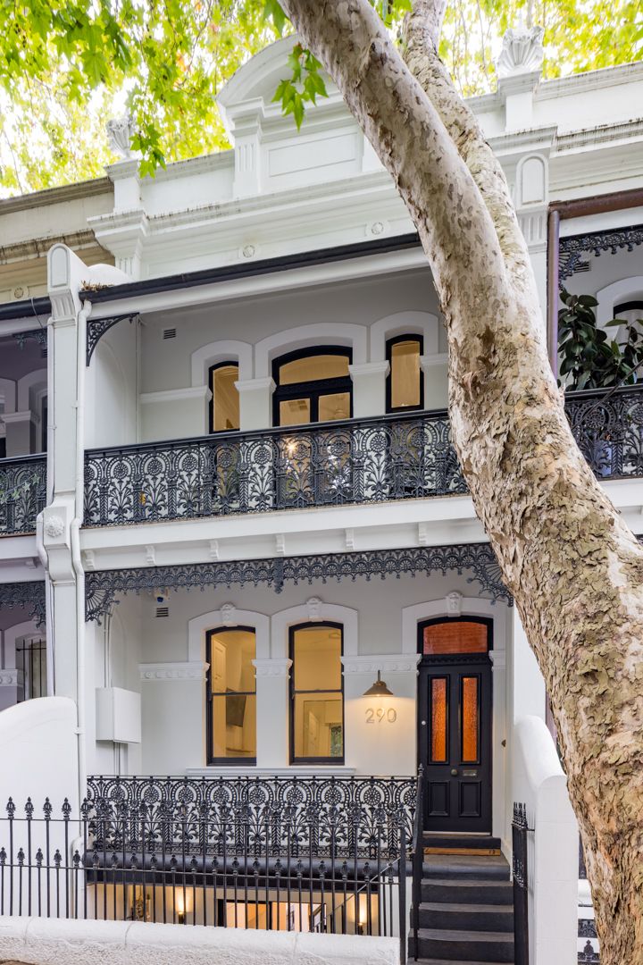 Victorian terrace with ornate black cast-iron lace balconies, arched windows and a dark front door behind a large tree trunk.