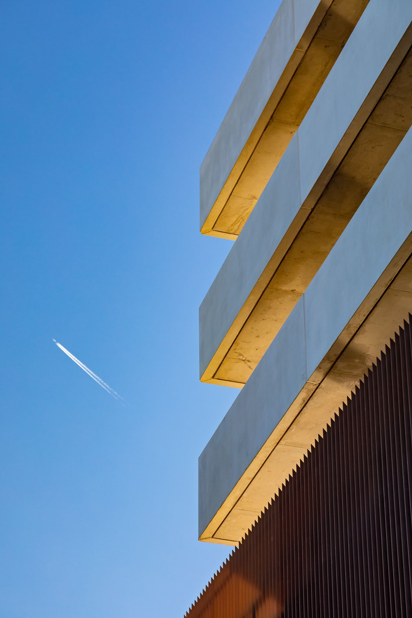 Stacked concrete building balconies with golden undersides beside a rusty slatted facade and a jet contrail in the blue sky.