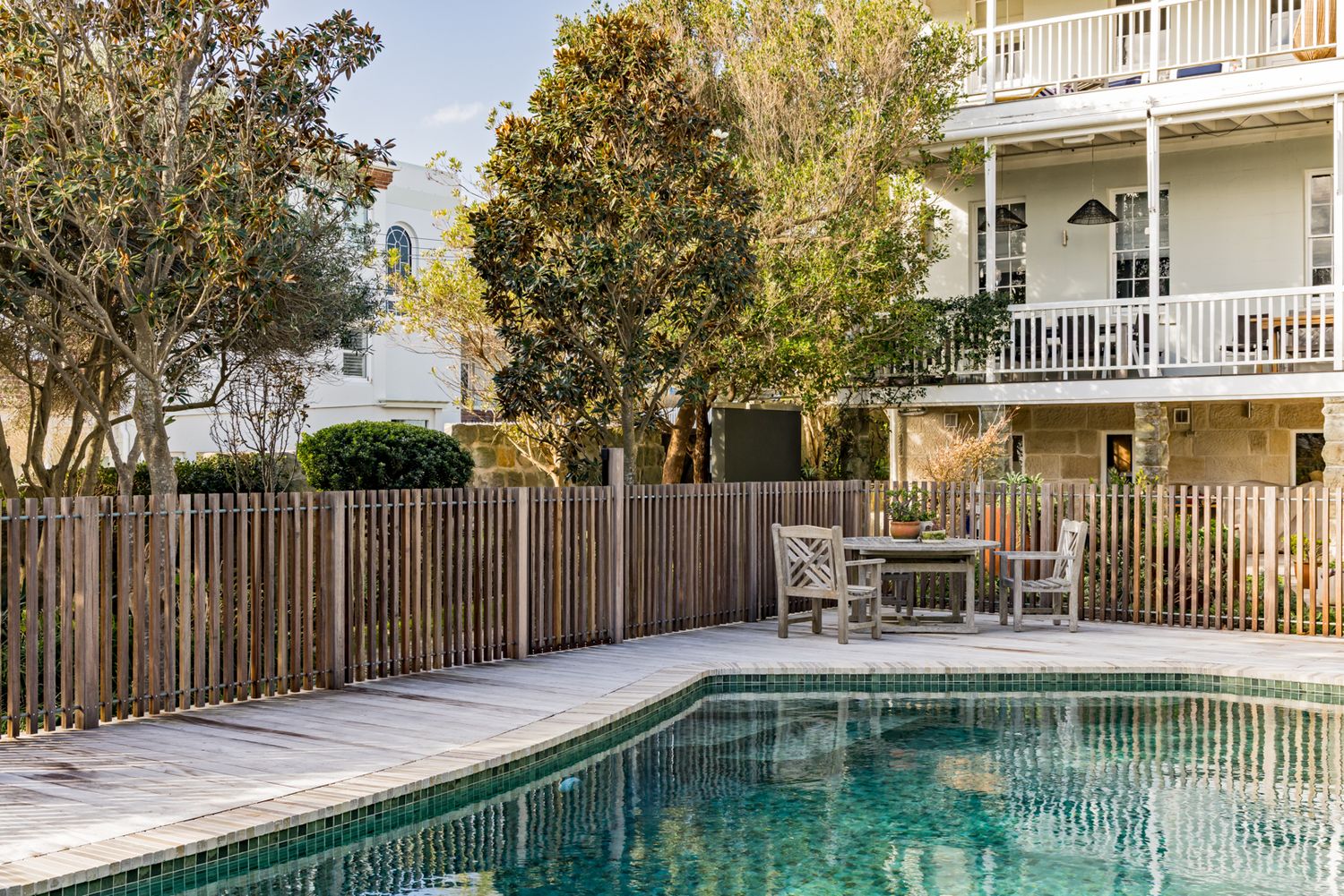 Swimming pool with clear green water beside a wooden fence and outdoor patio table, with a white multi-story building and trees behind.