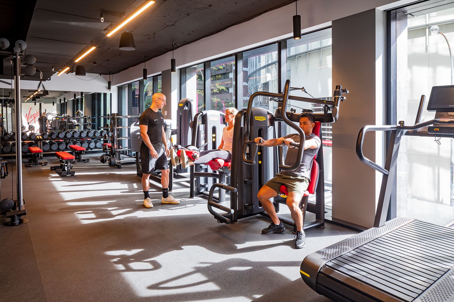 People exercise on strength machines in a spacious modern gym with large windows, track lighting and rows of cardio equipment.