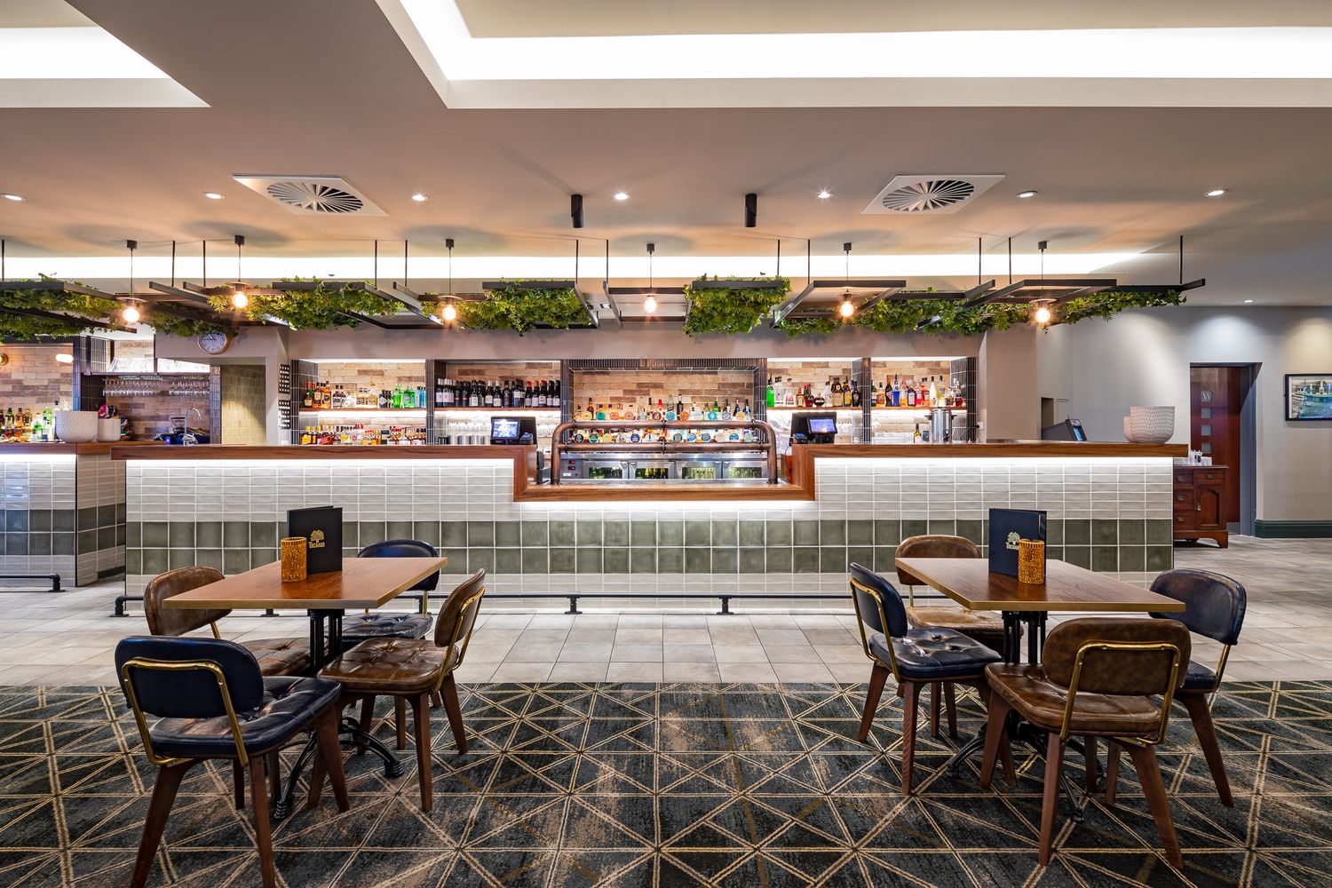 Modern restaurant interior with a green-tiled bar, hanging plants, wooden tables and chairs, and a geometric patterned floor.