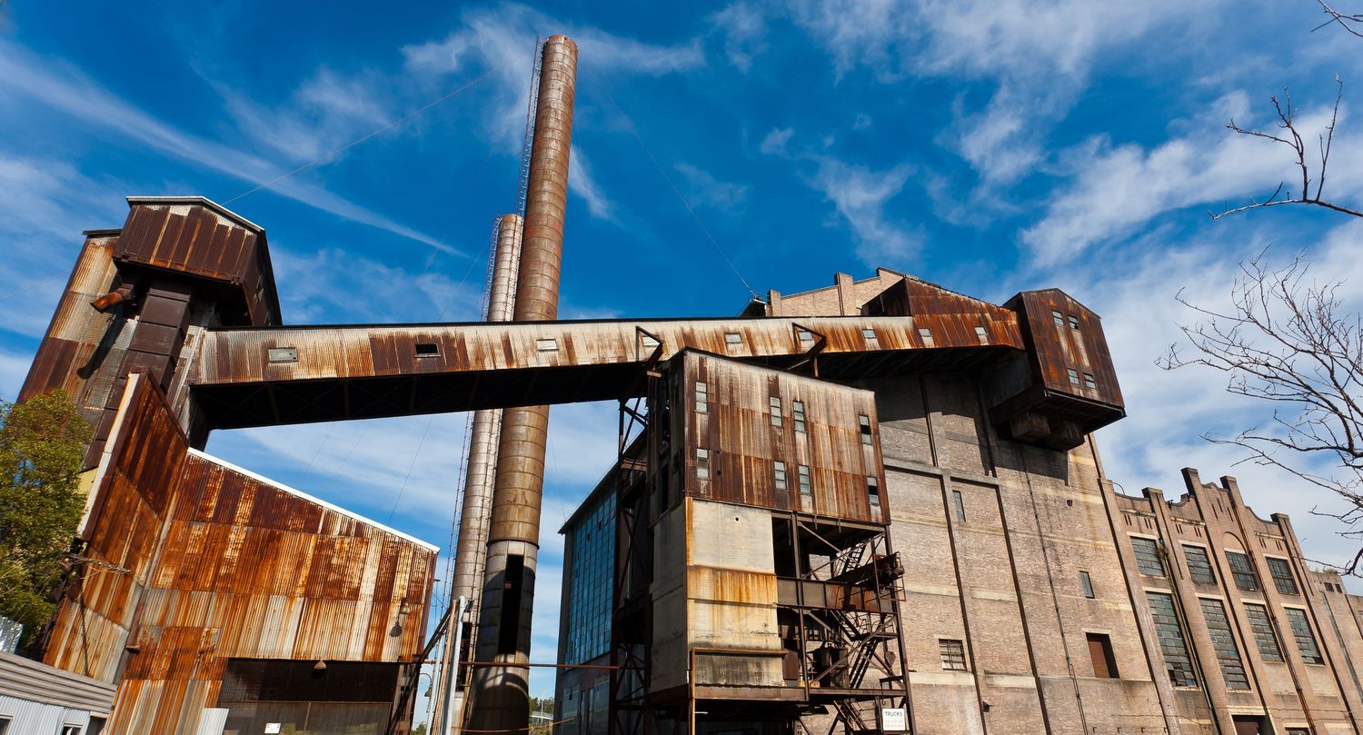 Rusted industrial ruins with tall brick chimneys and corroded metal structures set against a bright blue sky.