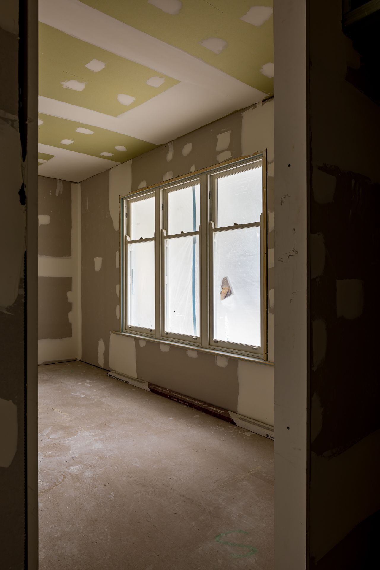 Empty room under renovation with bare plasterboard walls and ceiling and a large triple window letting in soft natural light.