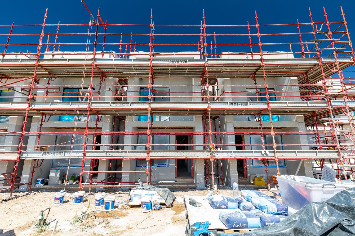 Townhouse building under construction wrapped in orange scaffolding against a blue sky, with building materials on the ground.