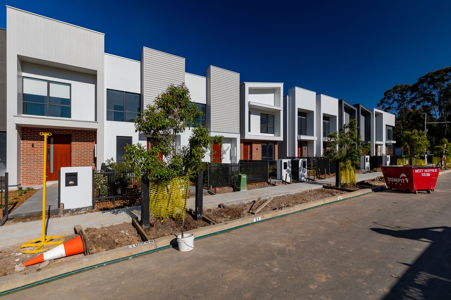 Modern townhouses with brick and white render facades, a red skip bin and traffic cones beside a freshly paved footpath.