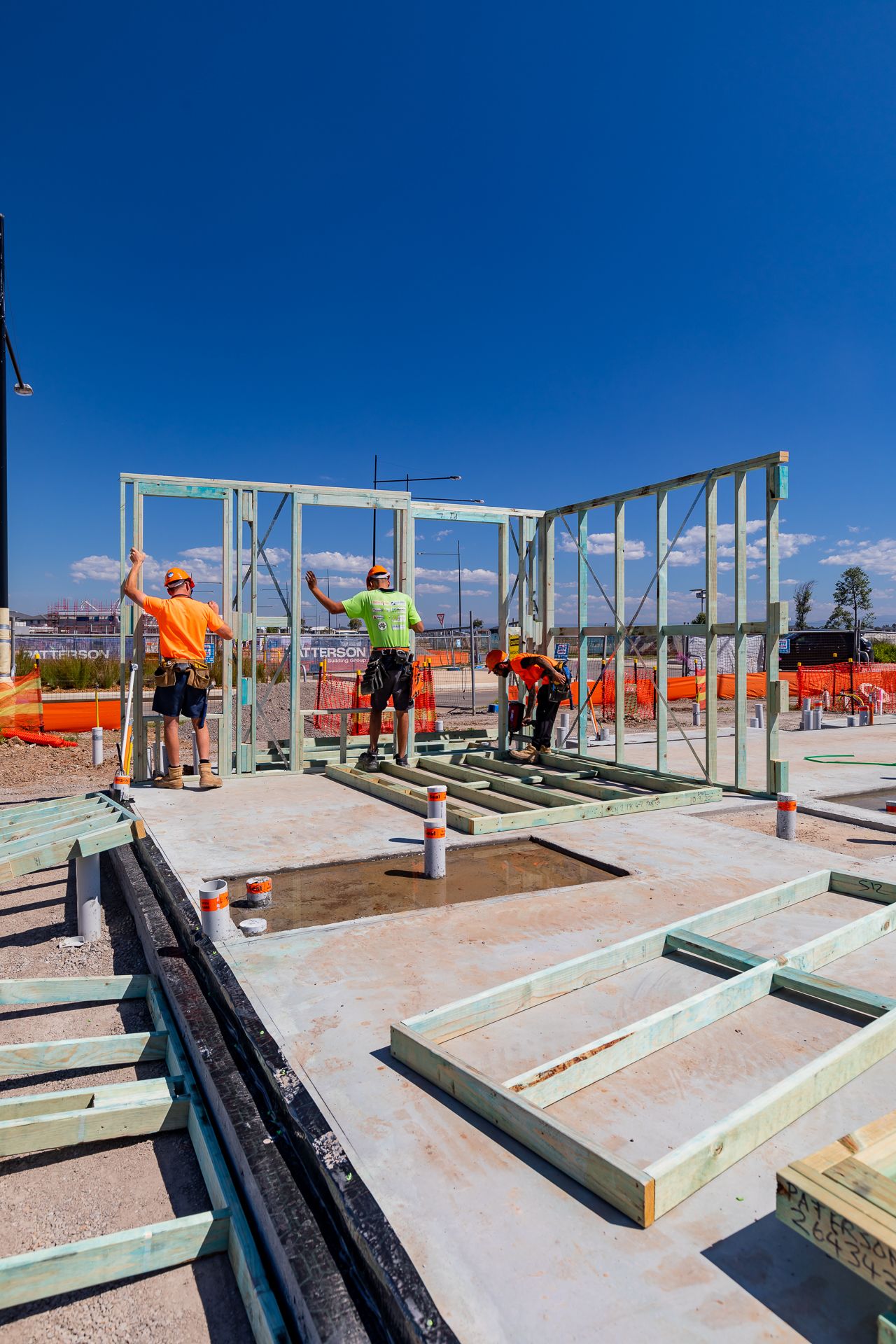 Construction workers in high-visibility shirts assembling a timber wall frame on a concrete slab under a clear blue sky.
