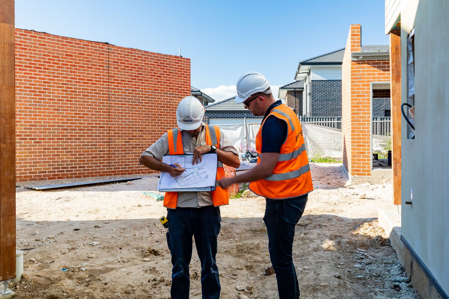 Two builders in hard hats and orange vests examine building plans on a site next to a new red brick wall.