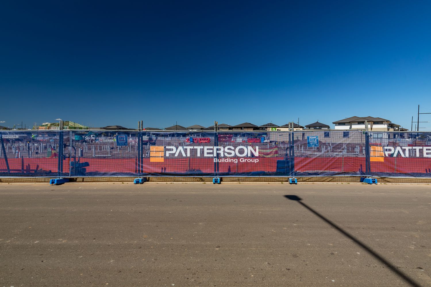 Patterson Building Group branded temporary fencing along a quiet road with houses visible behind under a deep blue sky.