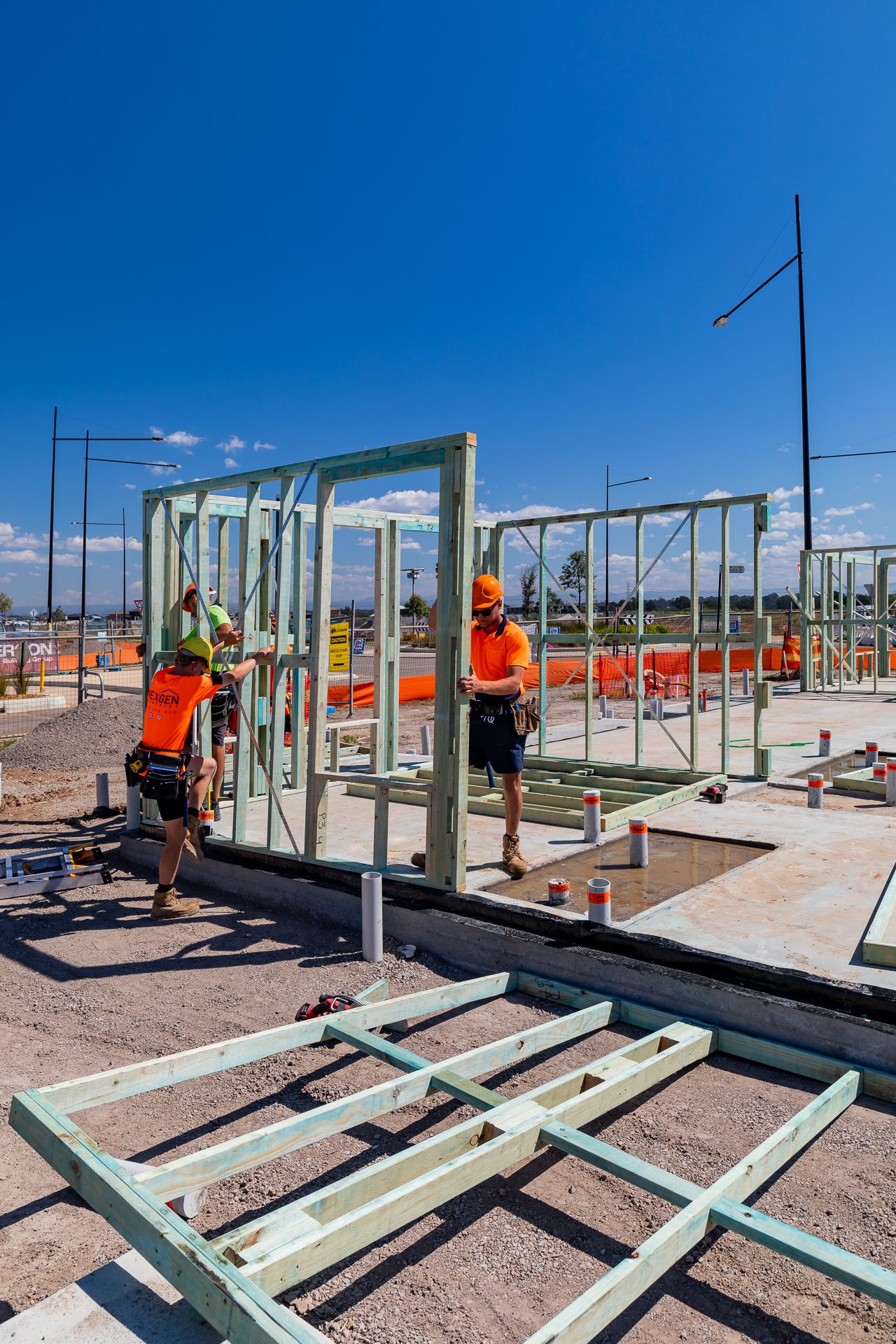 Builders in orange shirts and hard hats lift and position a steel wall frame on a house slab at a housing development.