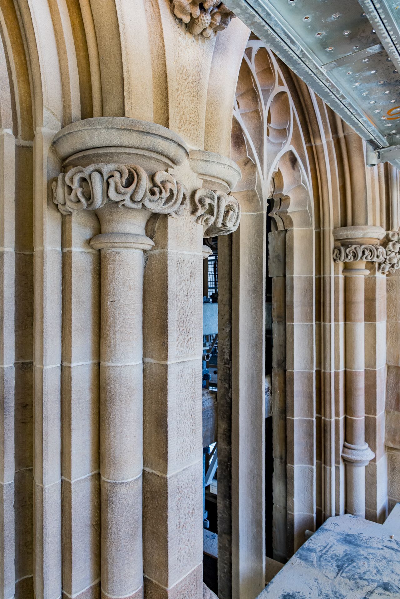 Ornate carved capital atop a sandstone pillar beside Gothic tracery windows in a historic building interior.