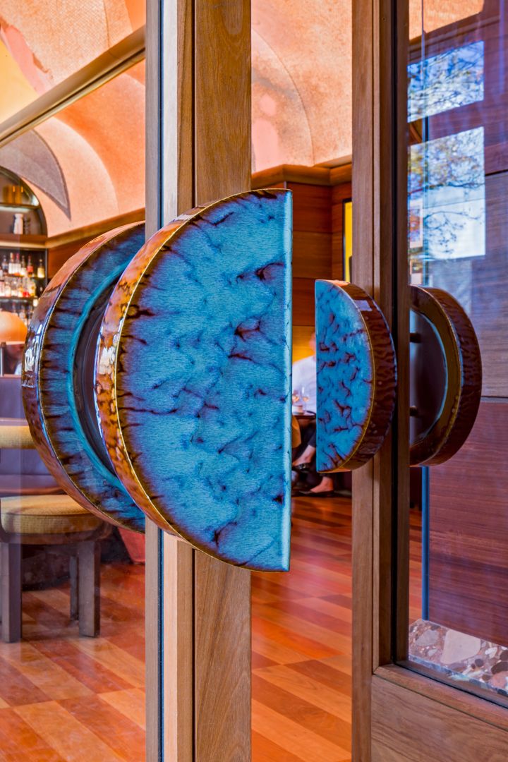 Close-up of oversized round blue ceramic door handles with a crackled glaze on a timber-framed glass door leading into a warm bar interior.