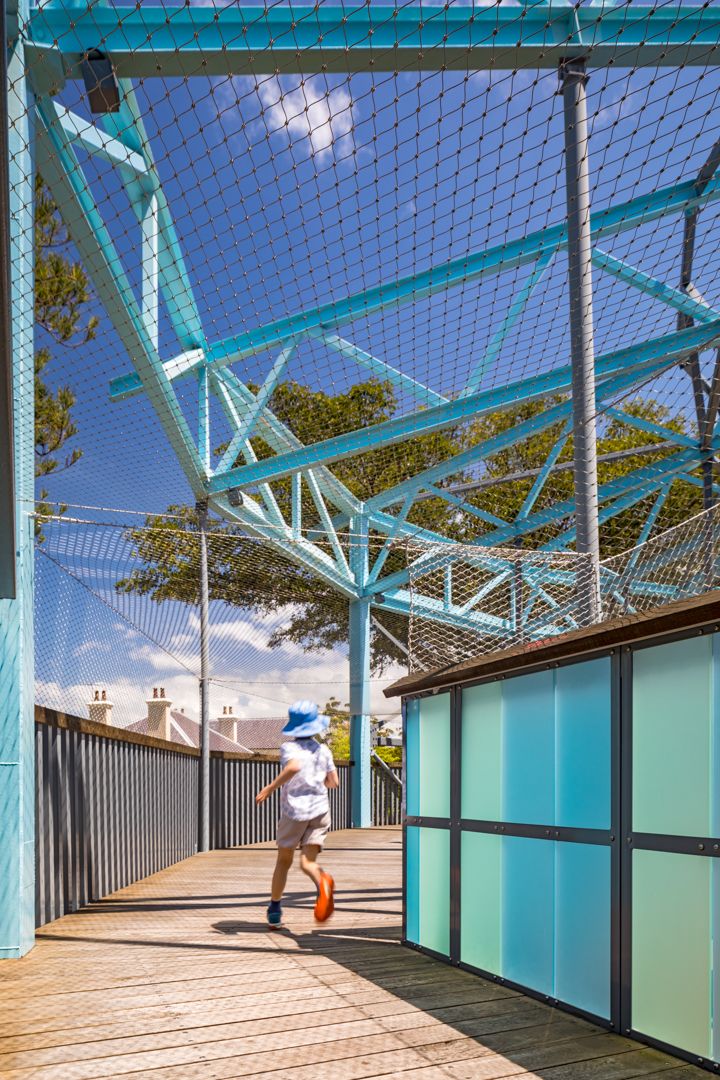 Child in a blue hat walks along a timber boardwalk beneath teal steel framework and mesh netting with turquoise panels.