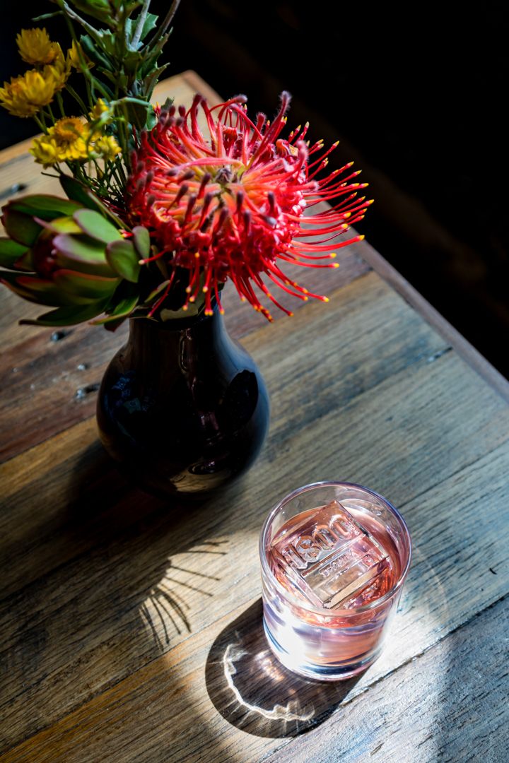 Red pincushion protea flower and yellow blooms in a dark vase on a rustic wooden table beside a pink cocktail with an engraved ice cube.