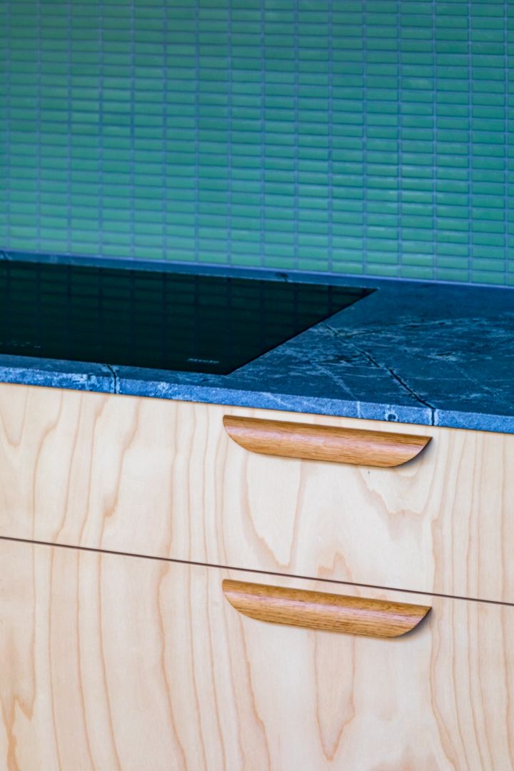 Light plywood kitchen drawers with curved oak handles beneath a dark stone countertop and green glazed tile backsplash.