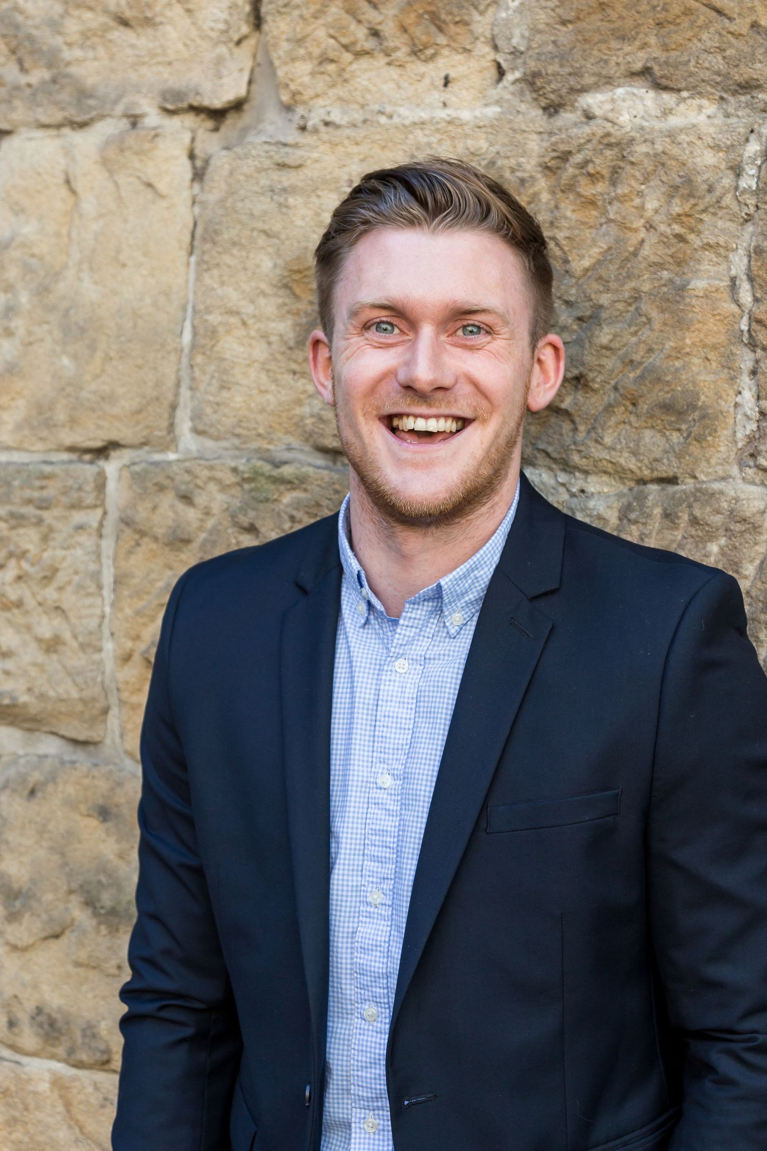 Laughing man with light stubble wearing a navy blazer and light blue checkered shirt standing in front of a sandstone wall.
