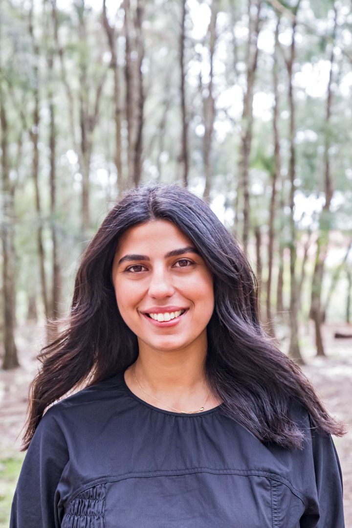 A smiling young woman with long dark hair and a black top stands in a sunlit forest of tall slender trees.